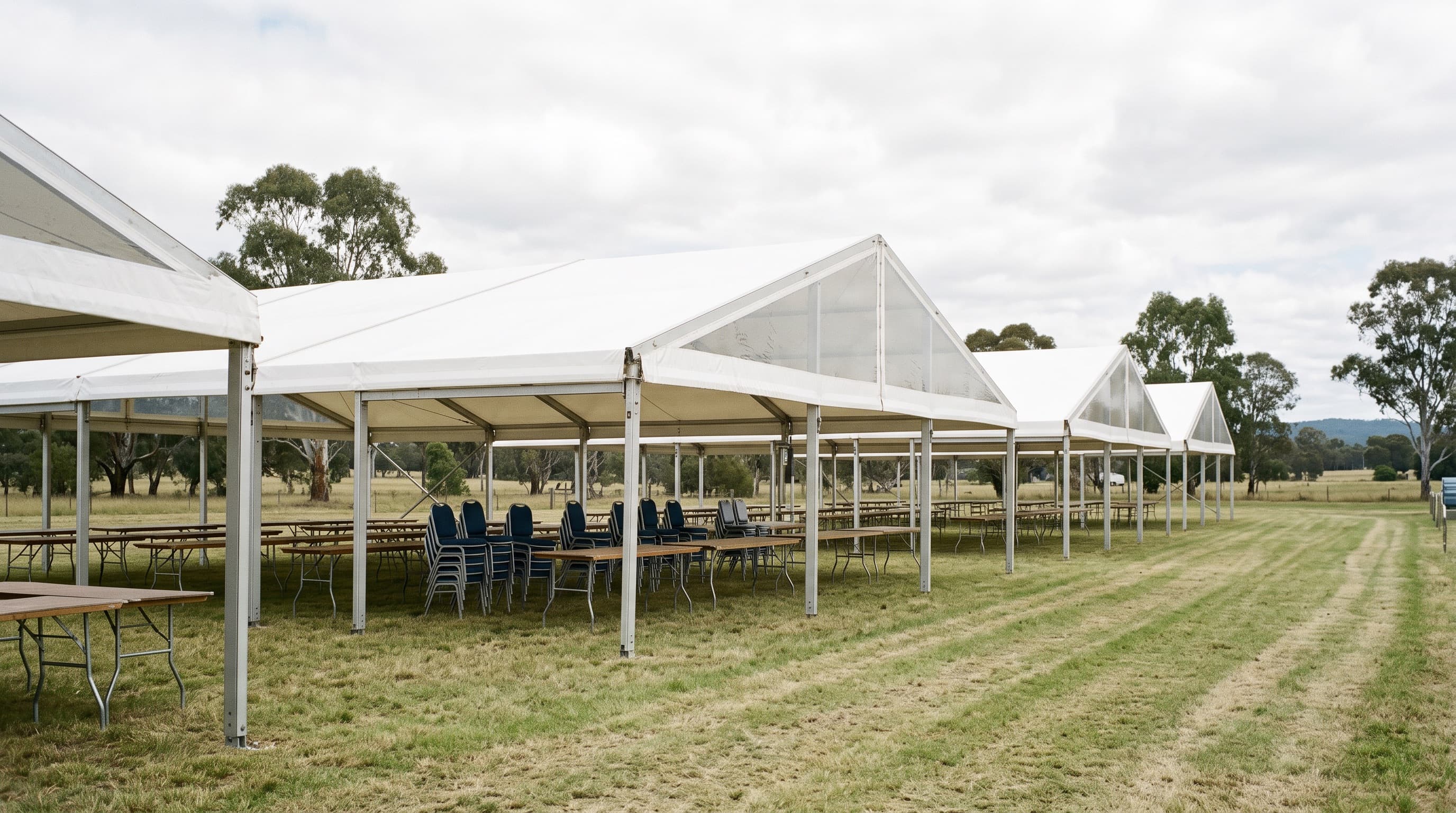 A run of linked white marquees with trestle tables and stacked chairs, set up in a paddock