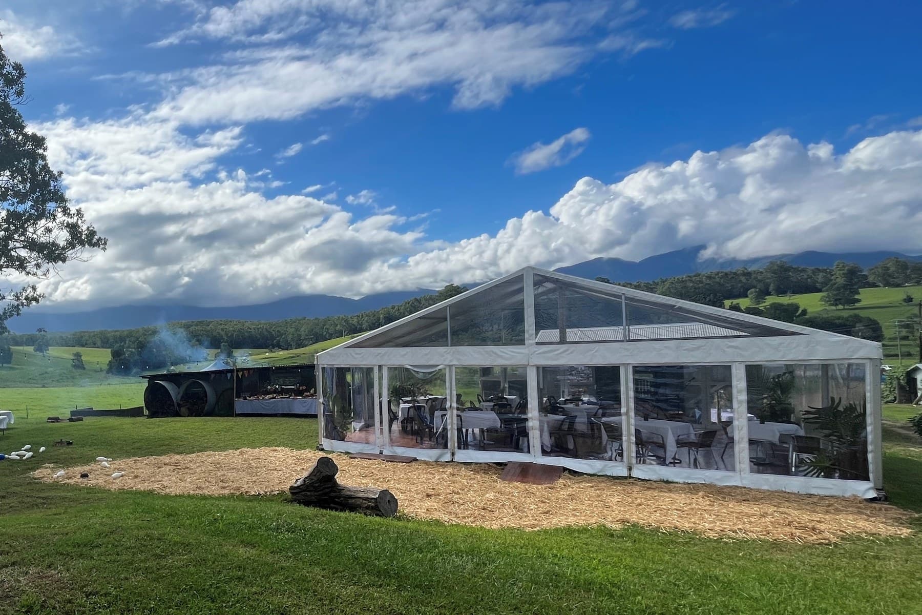 Clear-wall marquee set with tables for a seated dinner on a farm lawn below mountains