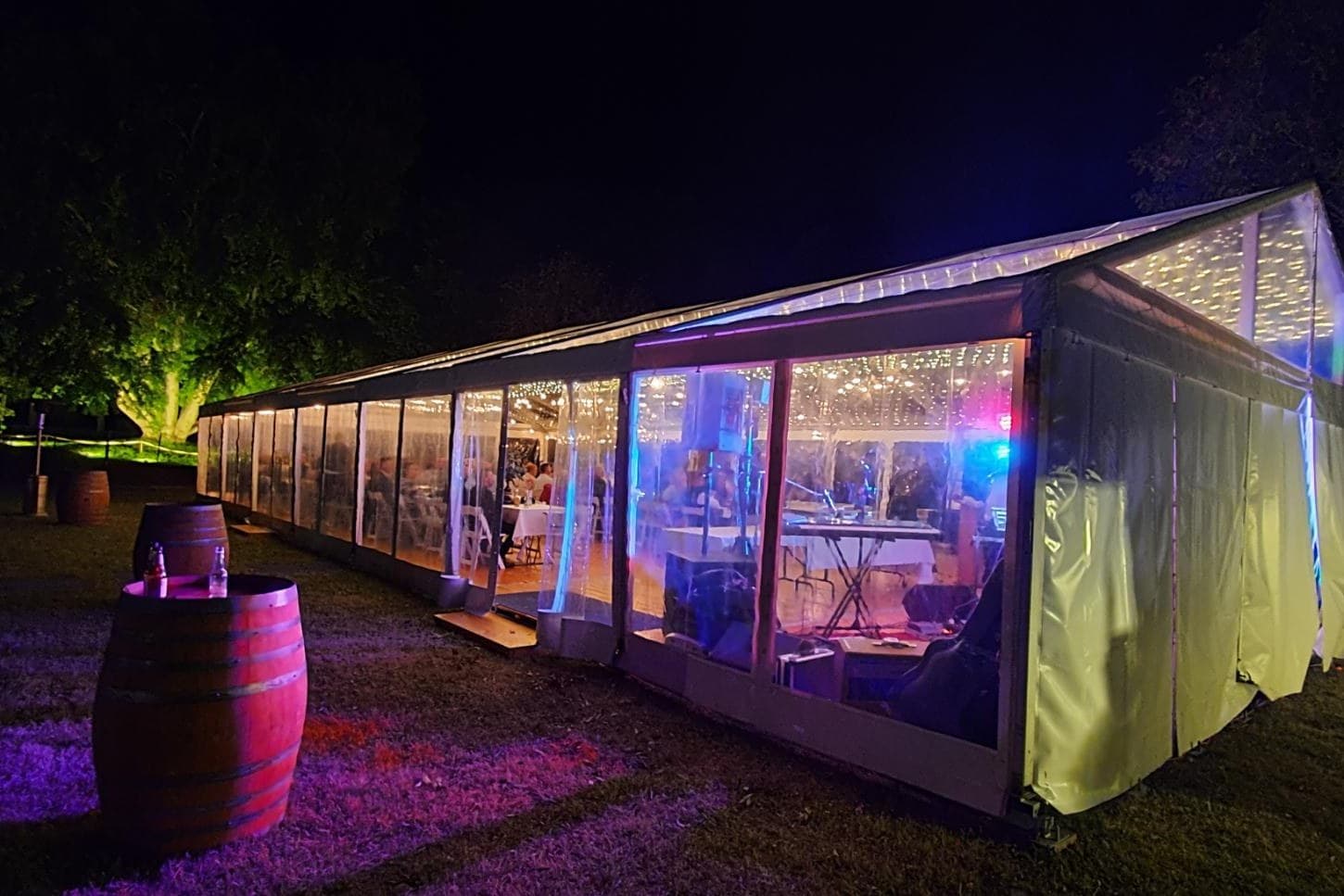 Clear-wall marquee lit with uplights at night, wine barrel on the lawn outside