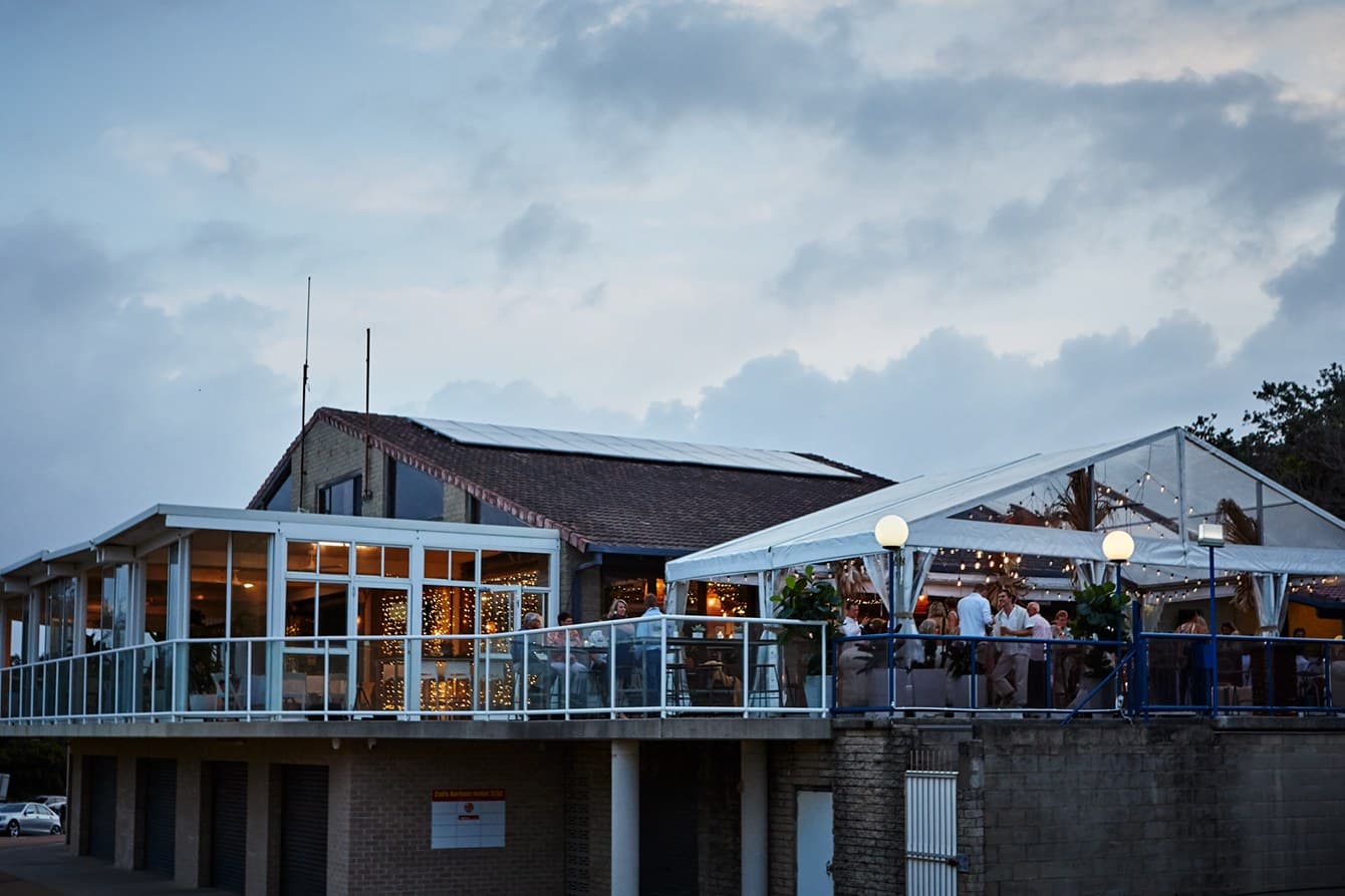 Clear-roof marquee on a surf club deck at dusk with guests seated inside